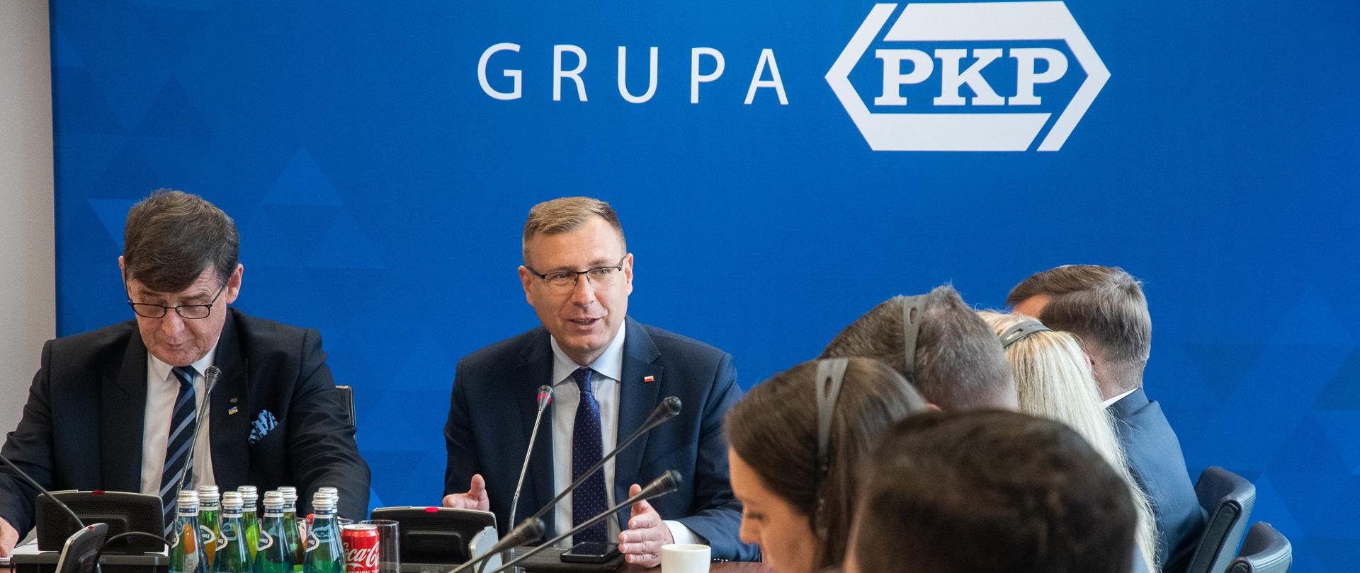 Wiceminister Maciej Małecki i prezes PKP S.A. Krzysztof Mamiński siedzą za stołem z innymi uczestnikami spotkania. W tle ścianka z logotypem Grupa PKP. 