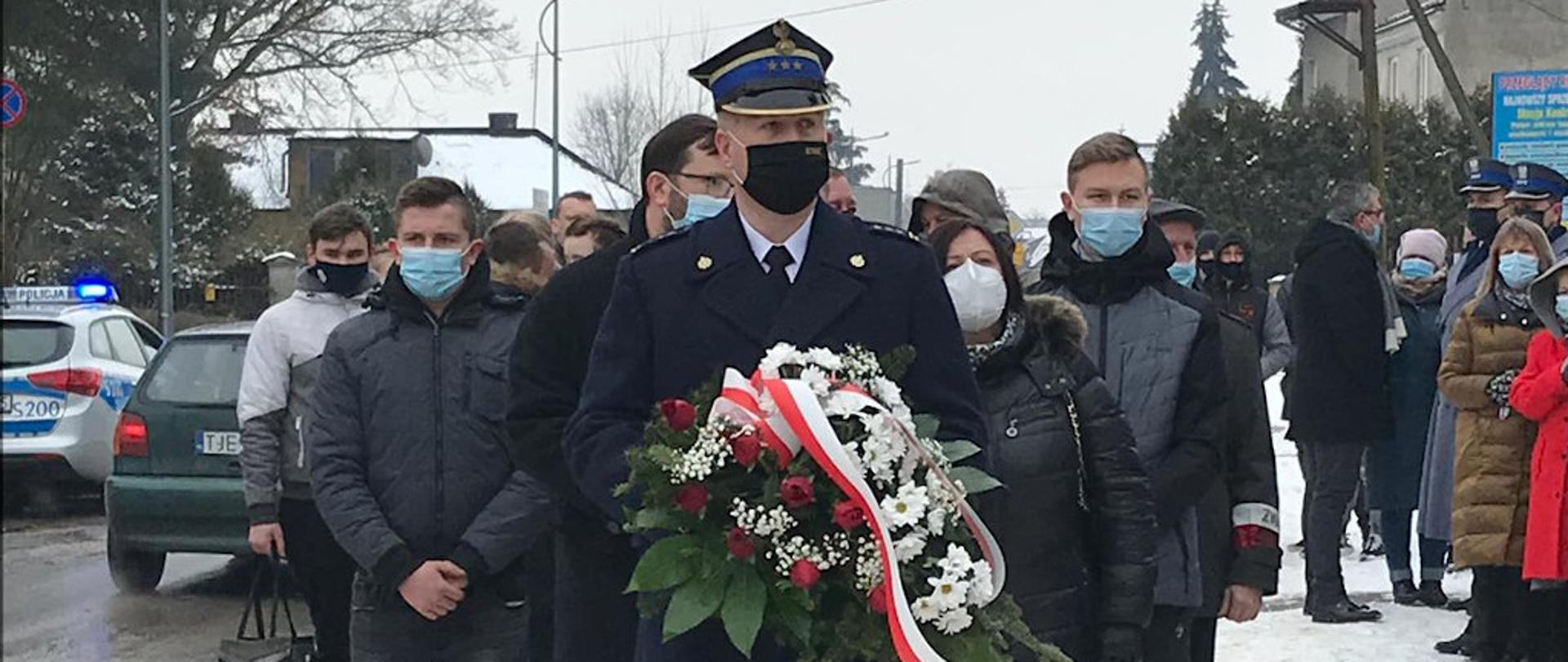 Na pierwszym palnie widnieje postać Komendanta Powiatowego Państwowej Straży Pożarnej w Jędrzejowie w umundurowaniu galowym oraz płaszczu. Na głowie ma założoną czapkę rogatywkę, natomiast na twarzy maseczkę ochronną zakrywającą usta i noc. W rękach trzyma wiązankę biało- czerwonych kwiatów. Za jego plecami znajdują się osoby czekające w kolejce do złożenia wiązanek kwiatów przed pomnikiem. Na twarzach mają założone maseczki ochronne.