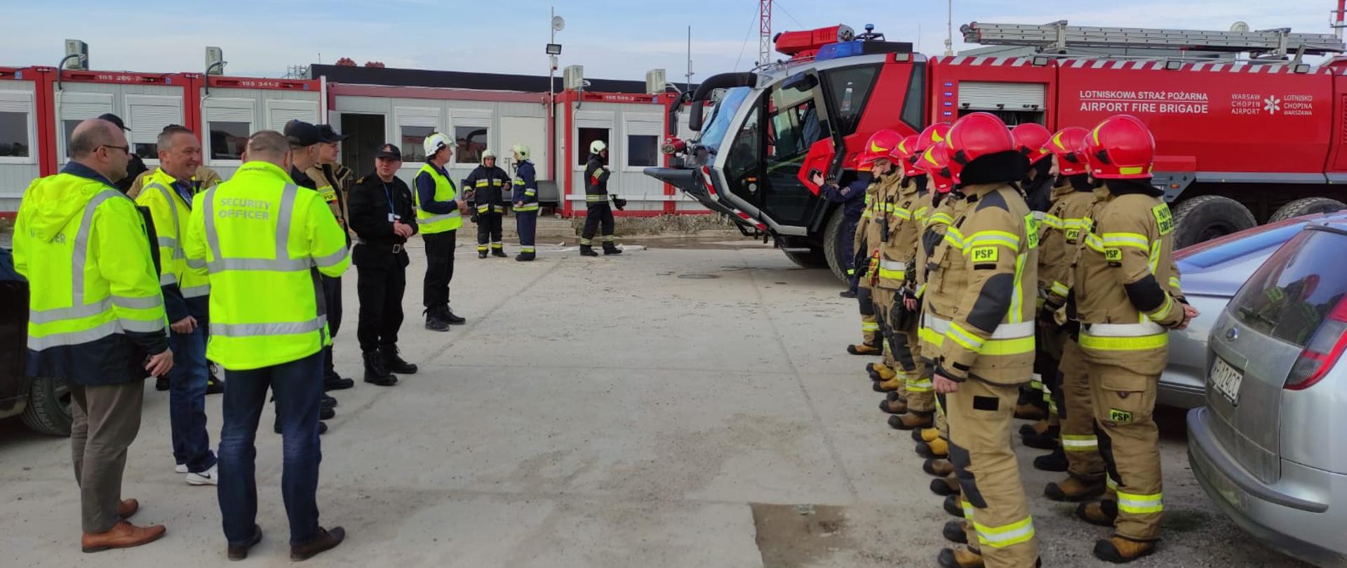 Zdjęcie przedstawia funkcjonariuszy Państwowej Straży Pożarnej, Lotniskowej Służby Ratowniczo - Gaśniczej oraz innych pracowników portu lotniczego. Strażacy ustawieni w dwu szeregu. frontem do nich stoją pracownicy portu oraz kadra kierownicza. trwa podsumowanie ćwiczeń. Strażacy są ubrani w ubrania specjalne w kolorze żółtym oraz ciemnym granatowym. Na głowach mają białe i czerwone hełmy. Kadra dowódcza ubrana głównie w ubrania dowódczo - sztabowe w kolorze czarnym. Pracownicy portu pozakładane mają kurtki i kamizelki w kolorze jasnym zielonym. W oddali po prawej stronie widać pojazd Lotniskowej Straży Pożarnej - Pantera. Pojazd w kolorze czerwonym ze srebrnymi elementami zabudowy. Kabina pojazdu jest przeszklona. Po lewej stronie w oddali znajdują się natomiast kontenery administracyjno - biurowe pracowników portu lotniczego. 