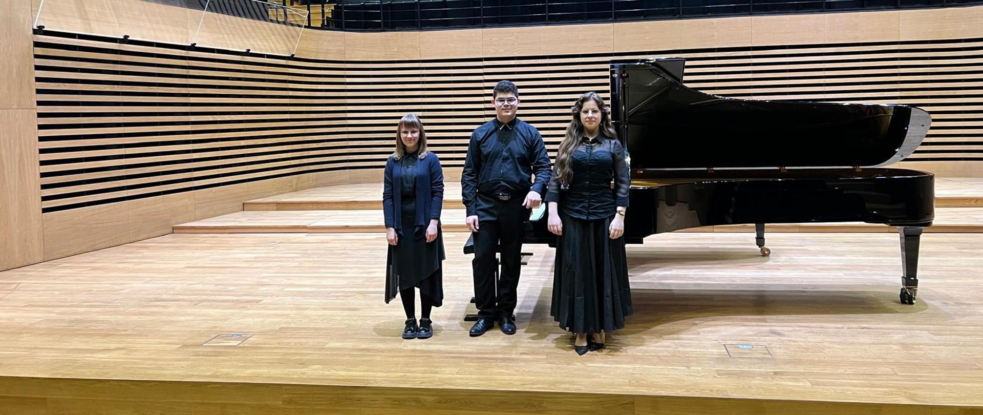 Dwie dziewczyny i chłopak stoją na scenie przed fortepianem.