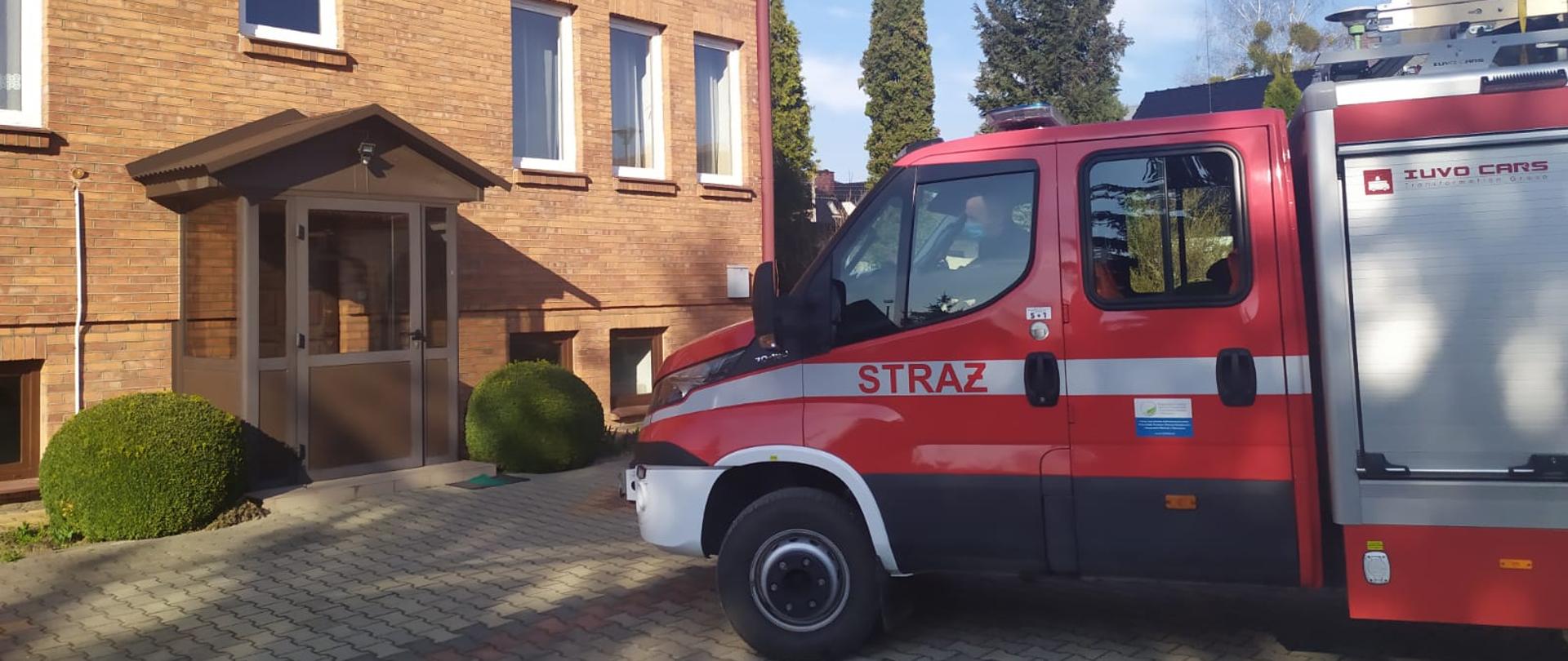 Zdjęcie przedstawia pojazd pożarniczy w kolorze czerwonym ze srebrnymi elementami zabudowy ustawiony przed budynkiem plebanii. Budynek dwupiętrowy, murowany z cegły, pokryty czerwoną blachą. 