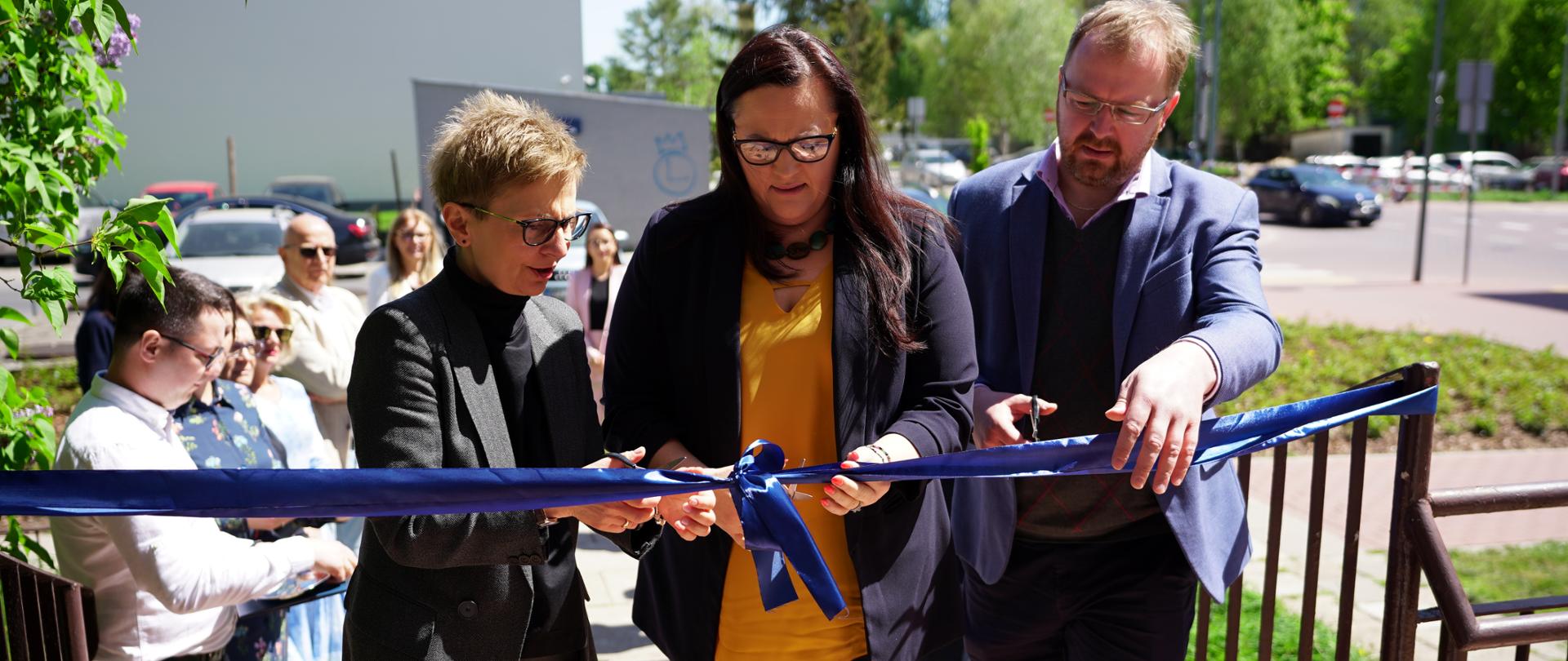 Trzy osoby przecinają wstęgę przed budynkiem. Druga od lewej wiceminister Małgorzata Jarosińska-Jedynak.
