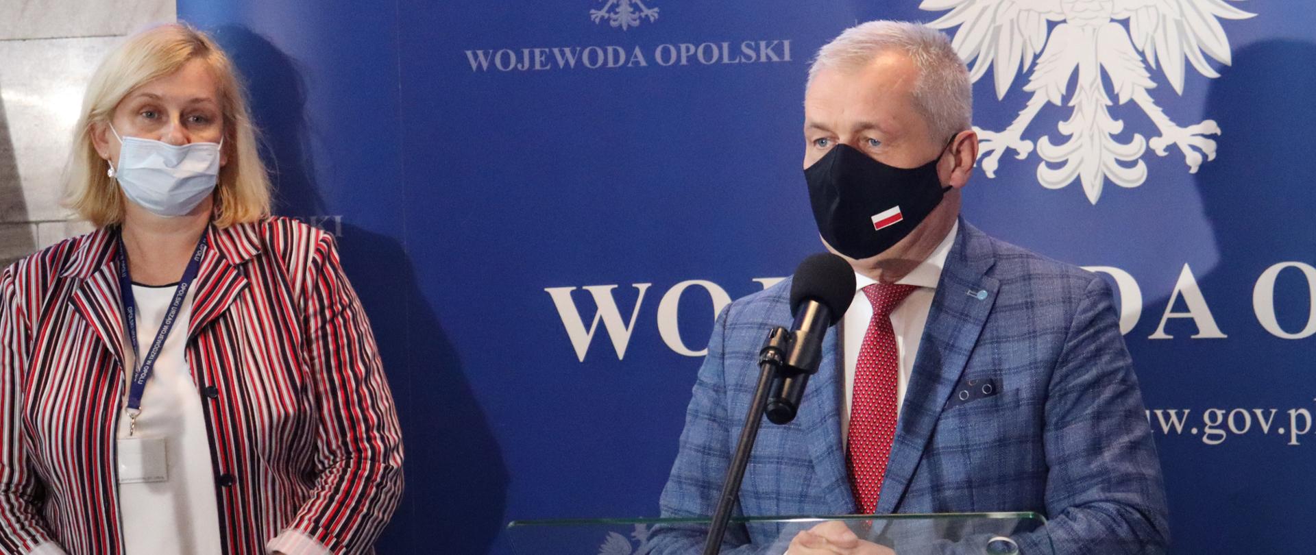 Na zdjęciu wojewoda opolski oraz dyrektorka Wydziału Zdrowia, przemawiają do mikrofonu podczas konferencji prasowej. Wojewoda stoi przy szklanej mównicy, po jego prawej stronie stoi dyrektorka wydziału. W tle granatowa ścianka z godłem Polski.