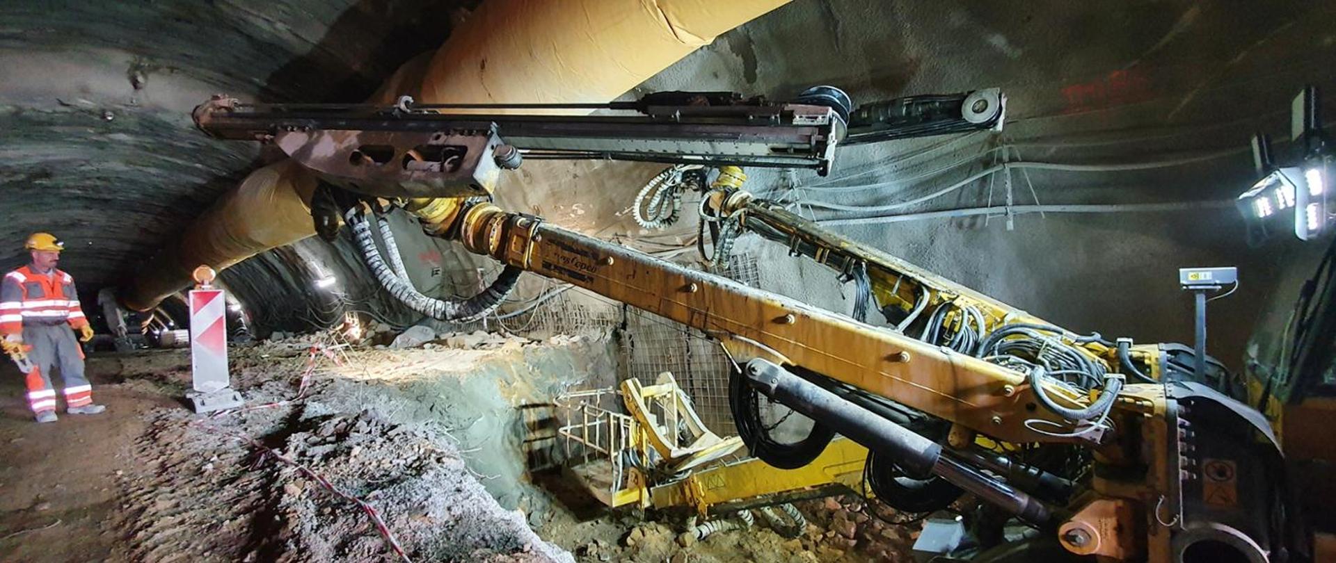Fotografia prac drążeniowych w tunelu. Widoczne maszyny specjalistyczne oraz pracownicy budowlani. Strop wydrążonej części zabezpieczony torkretem. 