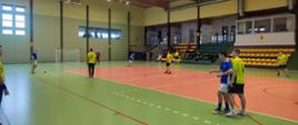Zawodnicy rywalizujący podczas meczu futsalu w hali sportowej – drużyny w żółtych i niebieskich strojach są w trakcie dynamicznej akcji na boisku. W tle widoczne są trybuny z kibicami oraz sędzia obserwujący przebieg spotkania - ciąg dalszy akcji.