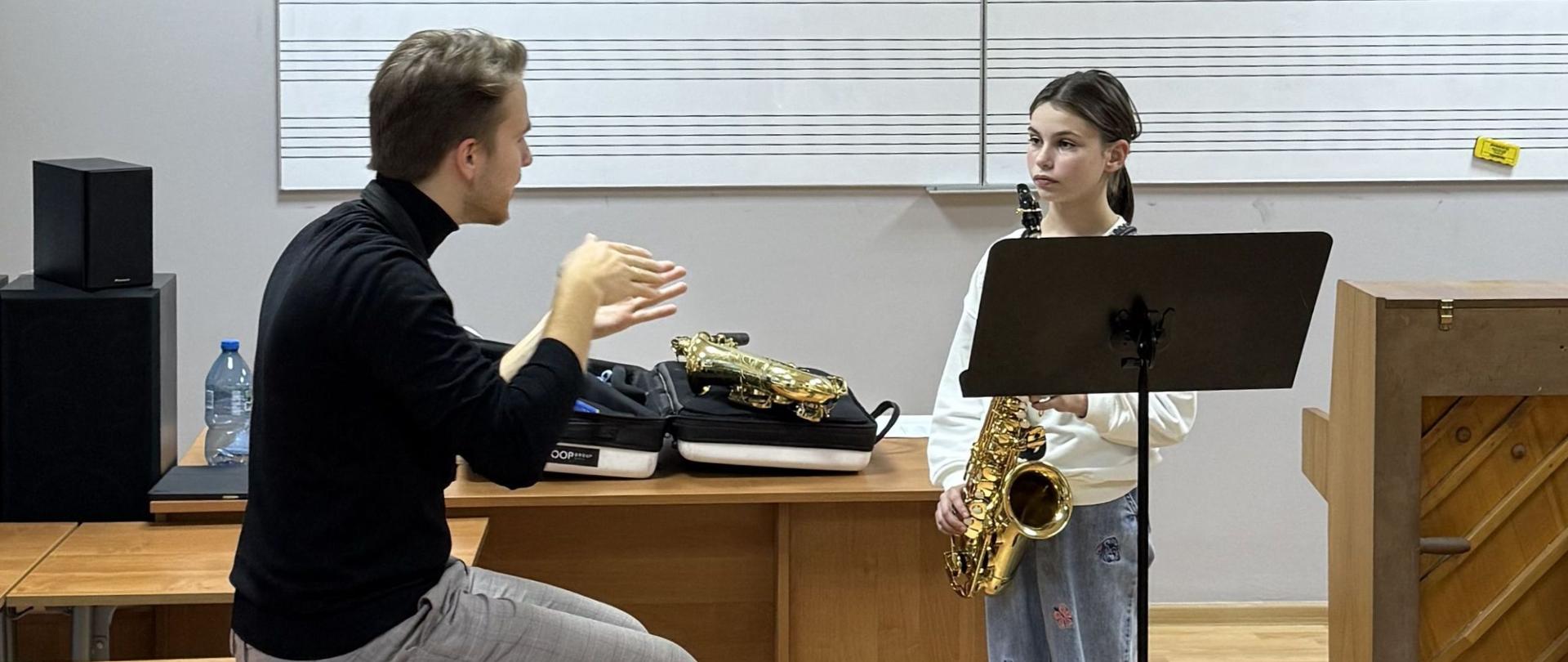 Anna Kędziora stoi trzymając saksofon. Słucha wskazówek pana Jakuba Murasa. Lekcja odbywa się w szkolnej sali. 