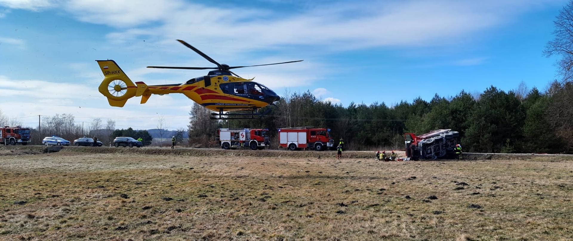 Zdjęcie przedstawia na pierwszym palnie lądujący śmigłowiec lotniczego pogotowia ratunkowego. Na drugim planie widnieją zaparkowane na jezdni samochody ratowniczo- gaśnicze z włączonymi błyskowymi światłami alarmowymi w kolorze niebieskim oraz wywrócona na prawy bok ciężarówka. Przed samochodem ciężarowym widać kilku strażaków, którzy udzielają kwalifikowanej pomocy medycznej kierującemu z samochodu ciężarowego. 