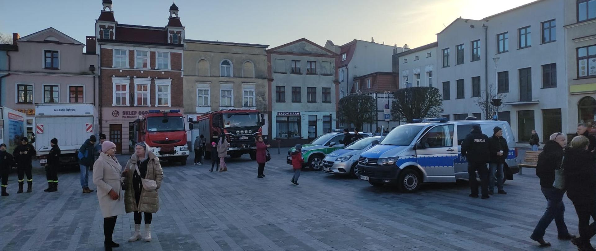 Na zdjęciu widać ustawione samochody służb mundurowych, które biorą udział w Finale WOŚP na rynku w Pucku, przy pojazdach znajdują się funkcjonariusze Straży Pożarnej, Policji oraz Straży Granicznej. 