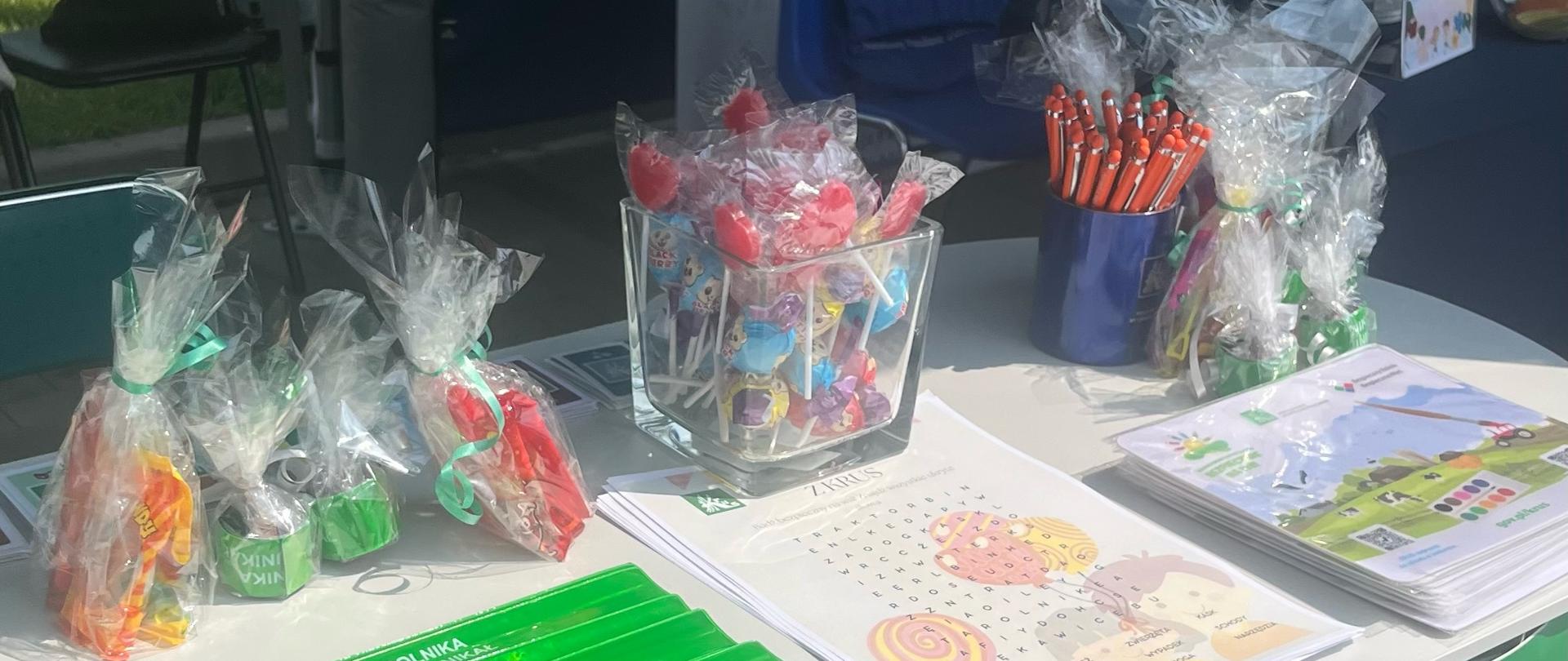 Stoisko promocyjne podczas obchodów Dnia Dziecka. Na stole znajdują się materiały edukacyjne oraz cukierki, lizaki, zielone opaski z napisem „Rola Rolnika”. Widoczna jest również tablica informacyjna z logo „Kasa Rolniczego Ubezpieczenia Społecznego” oraz banery „NFZ Narodowy Fundusz Zdrowia” i „Ministerstwo Zdrowia”.
