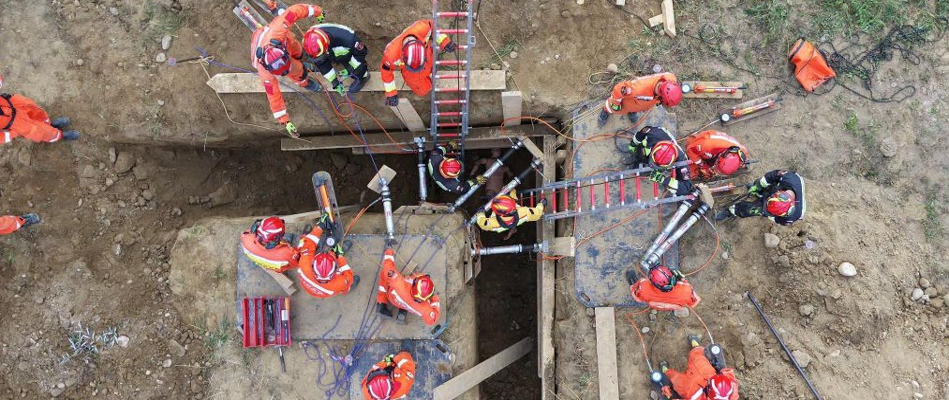 Zdjęcie z drona przedstawiające grupę kilkunastu strażaków w czerwonych mundurach i kaskach ochronnych, wykonujących ćwiczenia ratownicze w głębokim wykopie ziemnym. Scena widziana jest bezpośrednio z góry. Wykop ma nieregularny kształt z odnogami. Strażacy są rozmieszczeni wokół krawędzi oraz wewnątrz wykopu. Zabezpieczają ściany ziemne za pomocą drewnianych szalunków oraz specjalistycznych, srebrnych podpór hydraulicznych marki Holmatro. Widoczne są również liny ratownicze, drabiny oparte o ściany wykopu oraz żółty wąż wentylacyjny na dnie. Całość odbywa się na piaszczystym, otwartym terenie ćwiczeń.