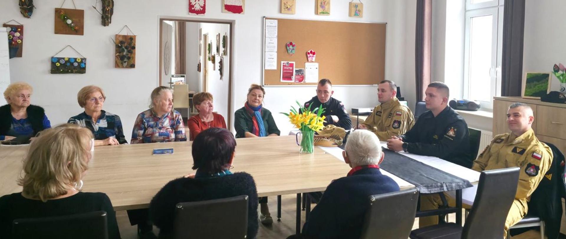 Zdjęcie przedstawia siedzących przy stole strażaków oraz seniorów. Zdjęcie wykonane w Akademii Bieszczadzkiego Seniora. 