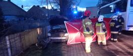Widać rozstawiony czerwony parawan, za nim uszkodzony srebrny samochód, przed parawanem jest czterech strażaków