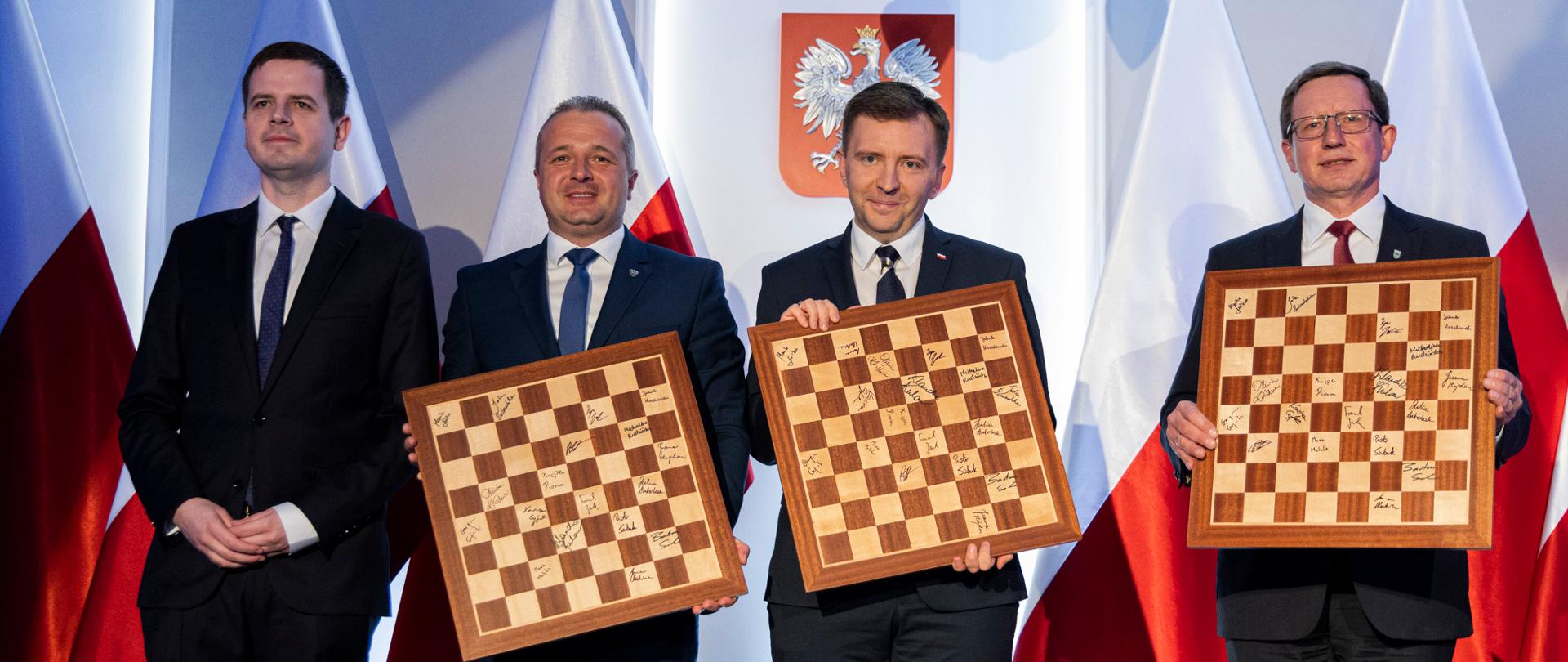 
4 osoby stojące z szachownicami w rękach na tle biało-czerwonych flag
