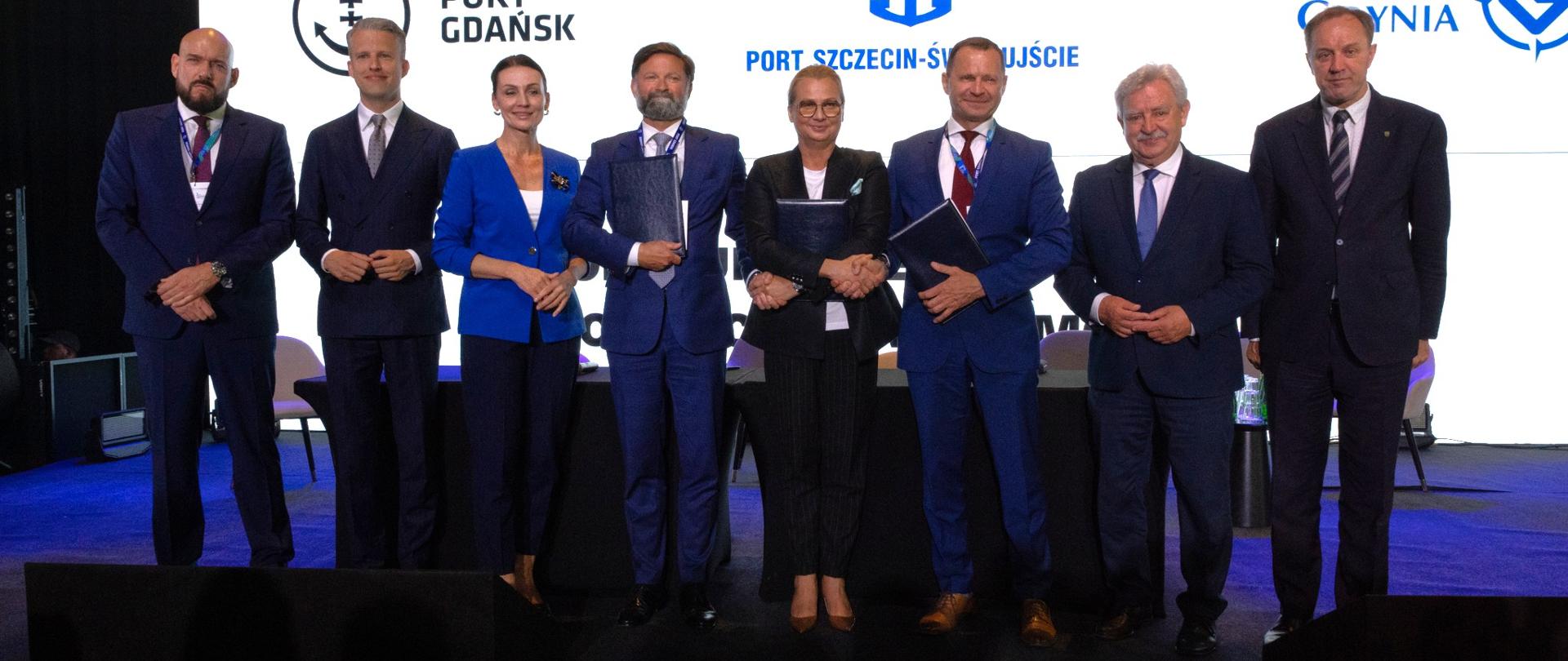 kilka osób trzyma podpisane umowy i pozuje do zdjęcia na tle tablicy z nazwami portów polskich