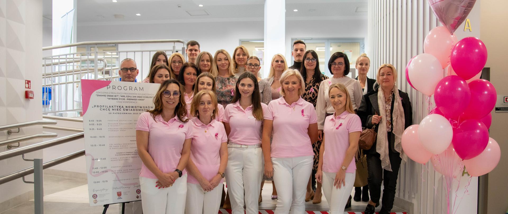 Na zdjęciu znajduje się grupa osób stojących na schodach. Po prawej stoją balony dekoracyjne w kolorze różowym, po lewej stronie plan konferencji. 