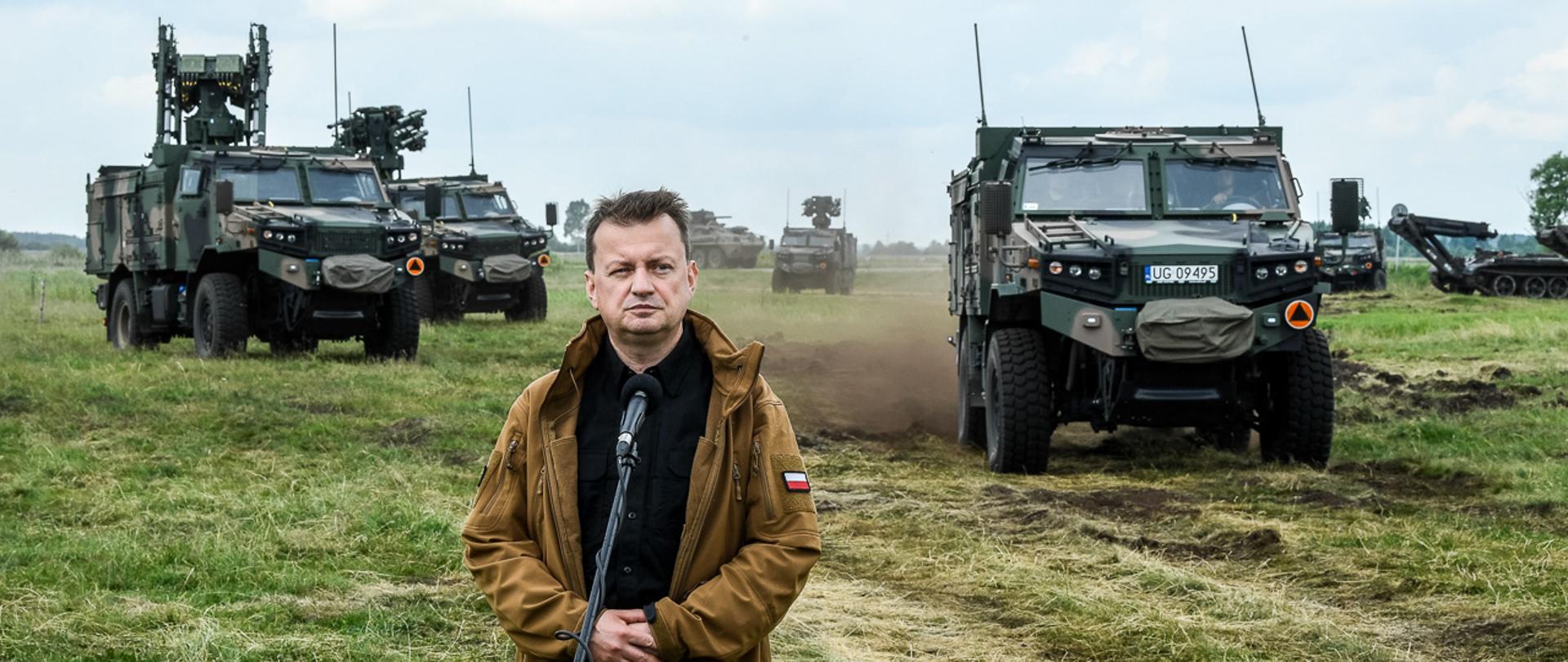 Szef MON przy mikrofonie podczas przemówienia na ćwiczeniach wojskowych. W tle pojazdy wojskowe.