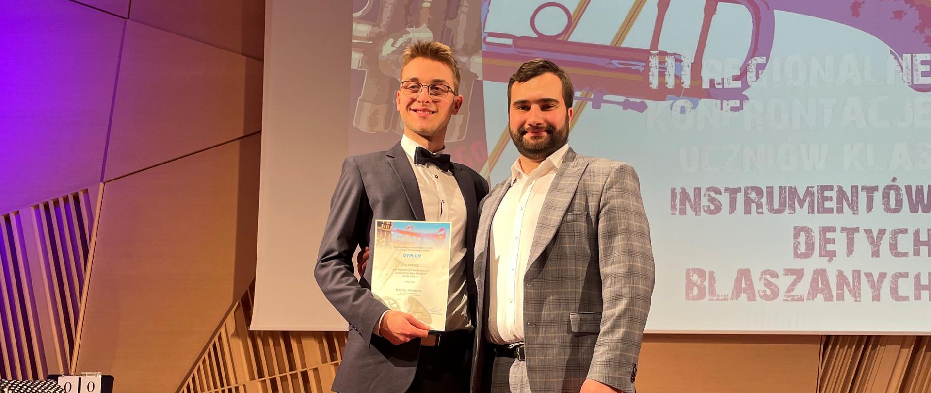 Zdjęcie przedstawia ucznia wraz z nauczycielem, którzy brali udział w konkursie "Instrumenty Dęte Blaszane" w Łodzi. Obaj panowie ubrani są w eleganckie garnitury.