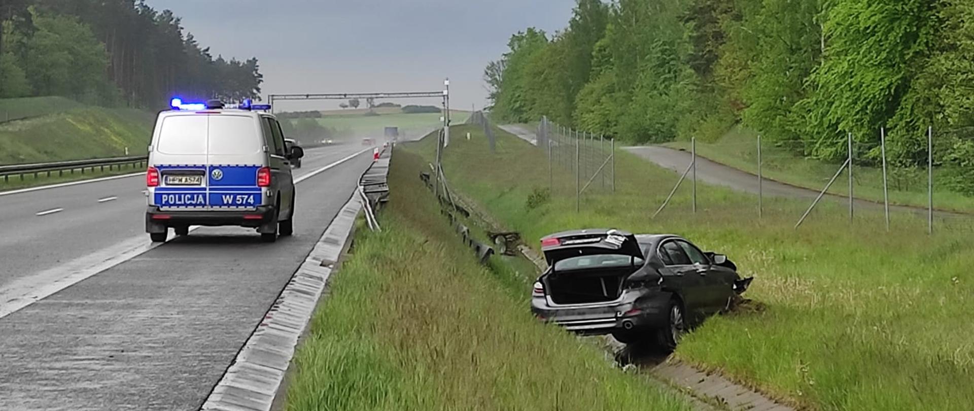 Na zdjęciu samochód osobowy leży w rowie przy drodze ekspresowej. Na pasie awaryjnym stoi radiowóz.