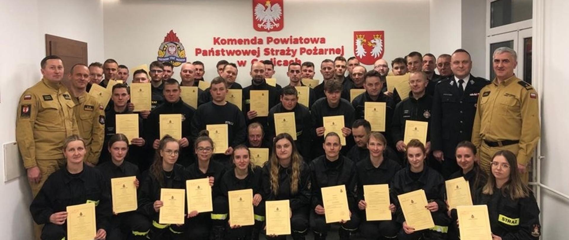 Na zdjęciu strażacy z Ochotniczych Straży Pożarnych ubrani w ubrania koszarowe koloru czarnego, zastępca komendanta powiatowego Państwowej Straży Pożarnej w Gorlicach i Prezes Zarządu Oddziału Powiatowego Związku Ochotniczych Straży Pożarnych Rzeczypospolitej Polskiej w Gorlicach oraz komisja egzaminacyjna ubrani w ubrania służbowe koloru piaskowego po egzaminie kończącym szkolenie podstawowe strażaków ratowników OSP w sali konferencyjnej w Komendzie Powiatowej Państwowej Straży Pożarnej w Gorlicach. 