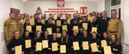 Na zdjęciu strażacy z Ochotniczych Straży Pożarnych ubrani w ubrania koszarowe koloru czarnego, zastępca komendanta powiatowego Państwowej Straży Pożarnej w Gorlicach i Prezes Zarządu Oddziału Powiatowego Związku Ochotniczych Straży Pożarnych Rzeczypospolitej Polskiej w Gorlicach oraz komisja egzaminacyjna ubrani w ubrania służbowe koloru piaskowego po egzaminie kończącym szkolenie podstawowe strażaków ratowników OSP w sali konferencyjnej w Komendzie Powiatowej Państwowej Straży Pożarnej w Gorlicach. 