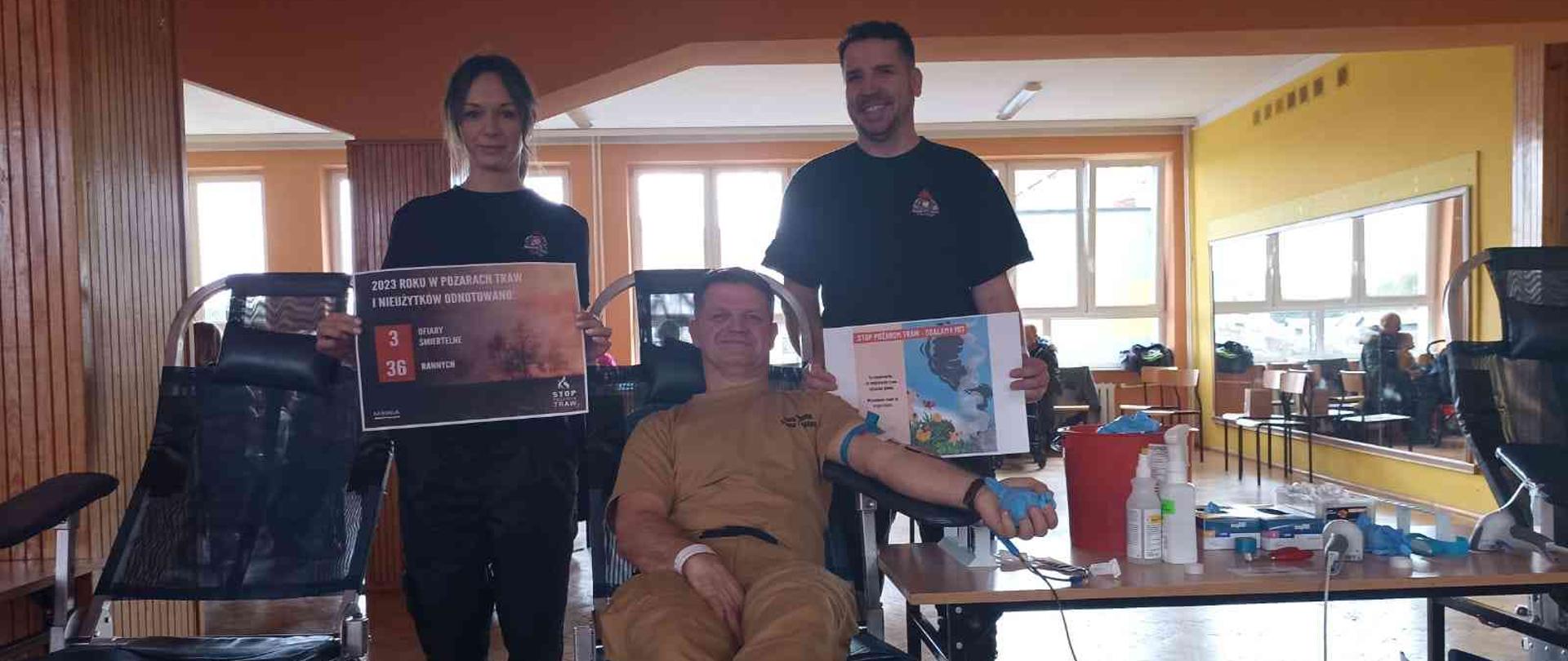 Strażak oddający krew, obok strażacy stojący z plakatami akcji.