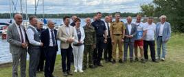 Na tle jeziora stoją przedstawiciele służb mundurowych i innych instytucji biorących udział w briefingu. 