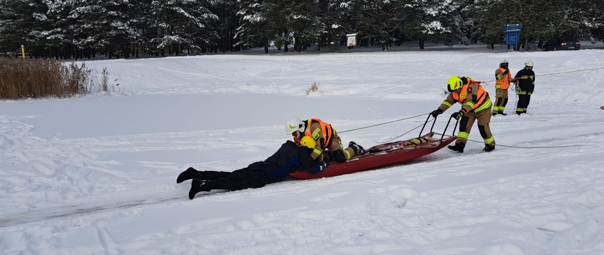 Na zaśnieżonym zbiorniku wodnym dwóch strażaków na saniach lodowych ewakuuje poszkodowanego.