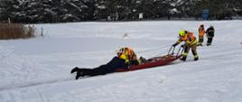 Na zaśnieżonym zbiorniku wodnym dwóch strażaków na saniach lodowych ewakuuje poszkodowanego.