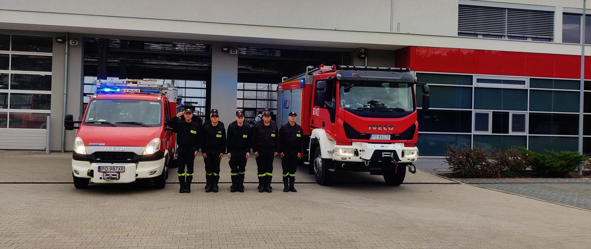Na zdjęciu stoją przed samochodami ratowniczo - gaśniczymi wystawionymi przed garaże JRG Śrem strażacy w dwu szeregu po lewej stronie stoi dowódca zmiany oddający honor 