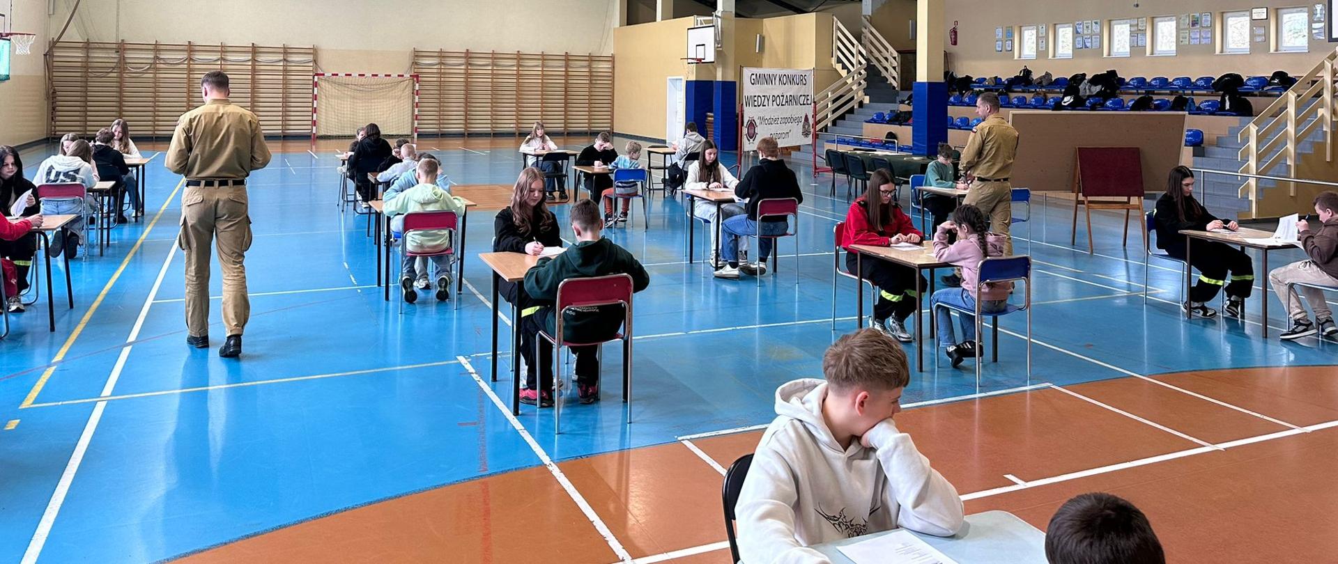 Na zdjęciu widać salę gimnastyczną w szkole. W środku ustawiono kilka stolików i krzeseł, przy których siedzą dzieci i młodzież. Każda osoba pracuje indywidualnie — większość pisze coś na kartkach, jest to ogólnopolski konkurs wiedzy pożarniczej na etapie gminnym. Wśród uczniów chodzą dwaj funkcjonariusze PSP kontrolując przebieg etapu pisemnego. 