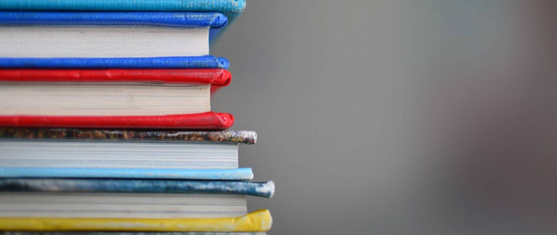 z lewej strony stos książek oprawionych w różnokolorowe okładki na szarym tle 