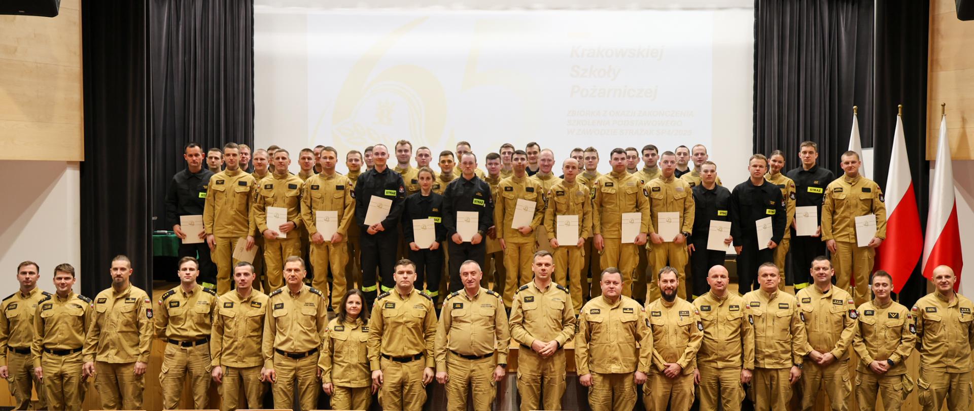 Cała grupa absolwentów i kadra stoi na scenie. Widoczne są rzędy strażaków trzymających w dłoniach certyfikaty. Jest to zdjęcie pamiątkowe z uroczystości zakończenia kursu, w tle widać flagi Polski.