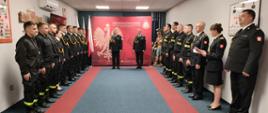 Zdjęcie przedstawia strażaków ustawionych w szeregach po obu stronach pomieszczenia. W środkowej części zdjęcia pomiędzy szeregami stoją Komendant Miejski oraz Dowódca Jednostki Ratowniczo-Gaśniczej.