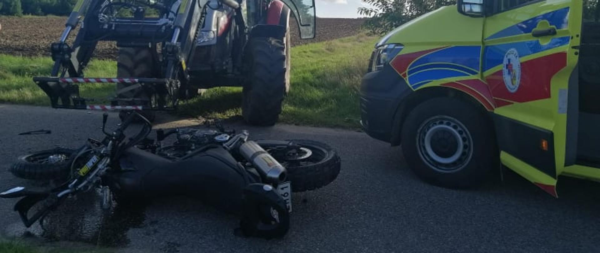 Motocykl leży przewrócony na bok na drodze. Ciągnik rolniczy stoi na wyjeździe z łąki. Przed nimi stoi karetka pogotowia.