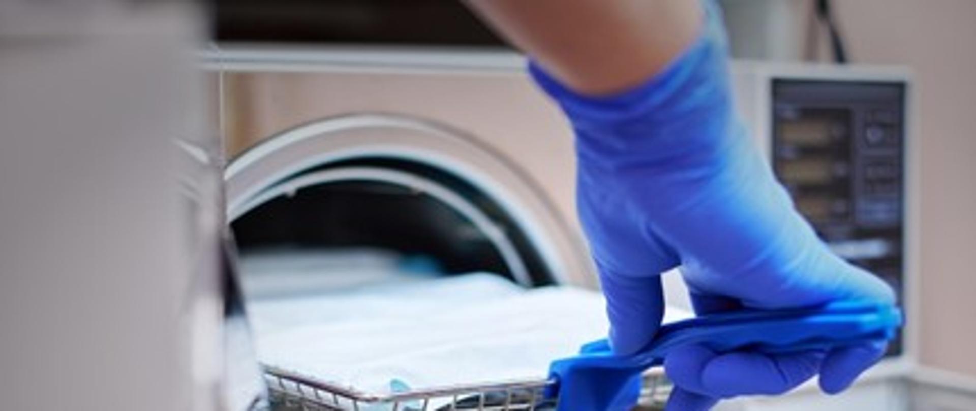 Na obrazku widać rękę pracownika medycznego ubraną w niebieską rękawiczkę jednorazową, który zamyka urządzenie do sterylizacji