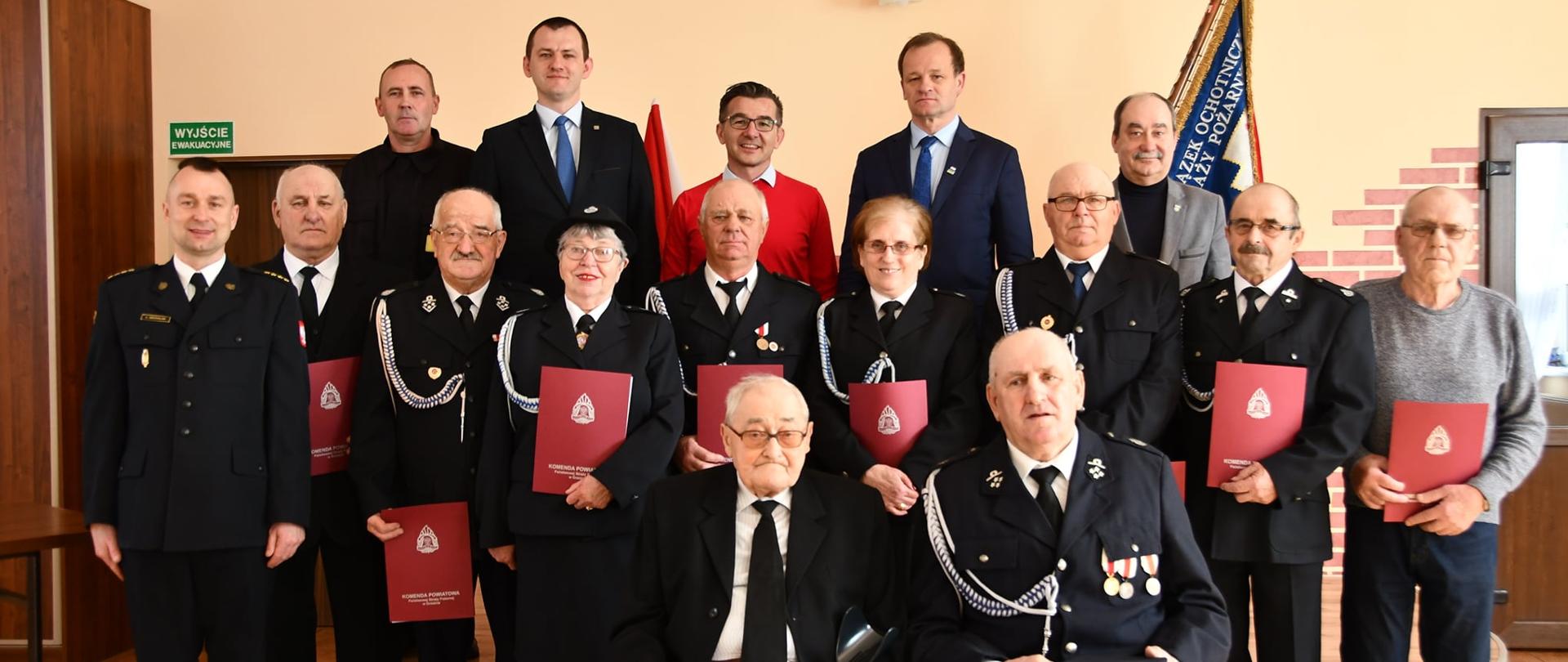 Zdjęcie grupowe uhonorowanych druhen i druhów wraz z osobami wręczającymi na tle sztandaru OSP