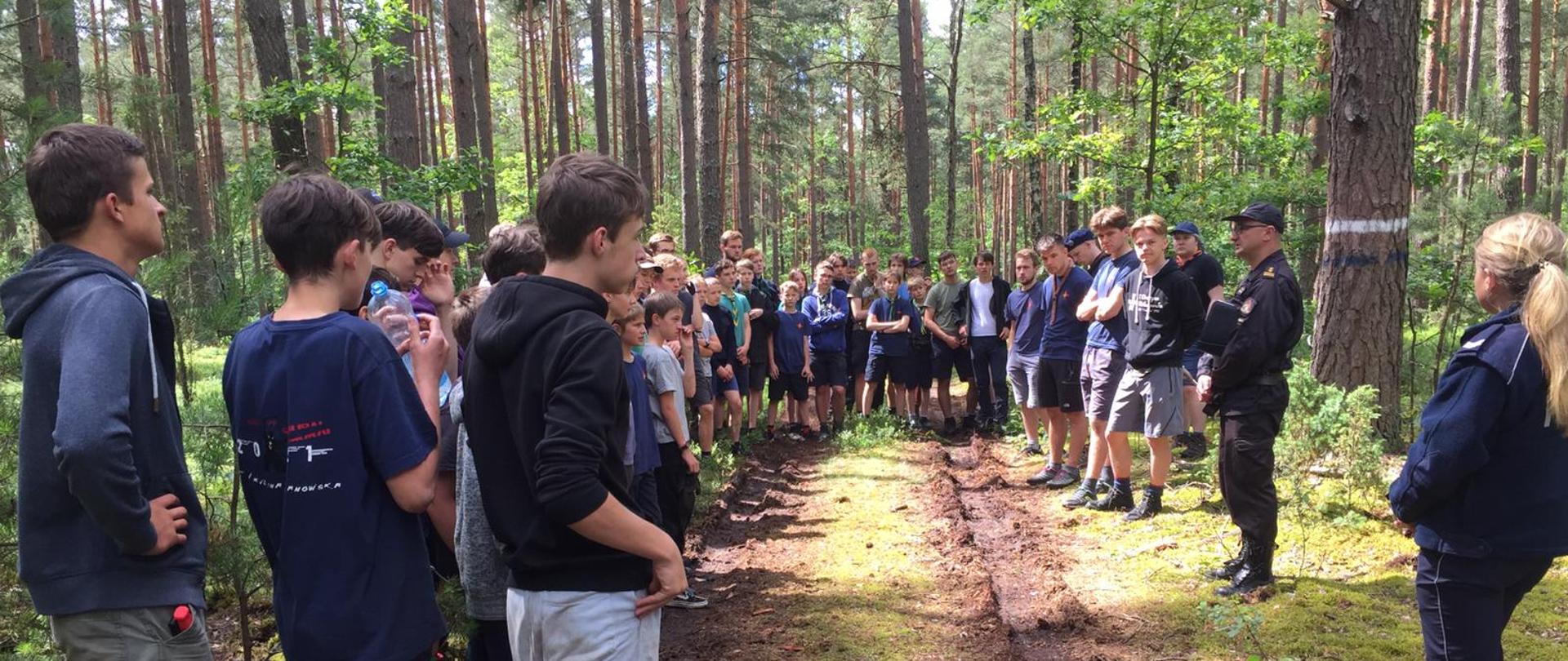 droga leśna, po lewej stronie i wgłąb harcerze w trakcie pogadanki którą przeprowadzają: strażak i funkcjonariuszka Policji stojący po prawej