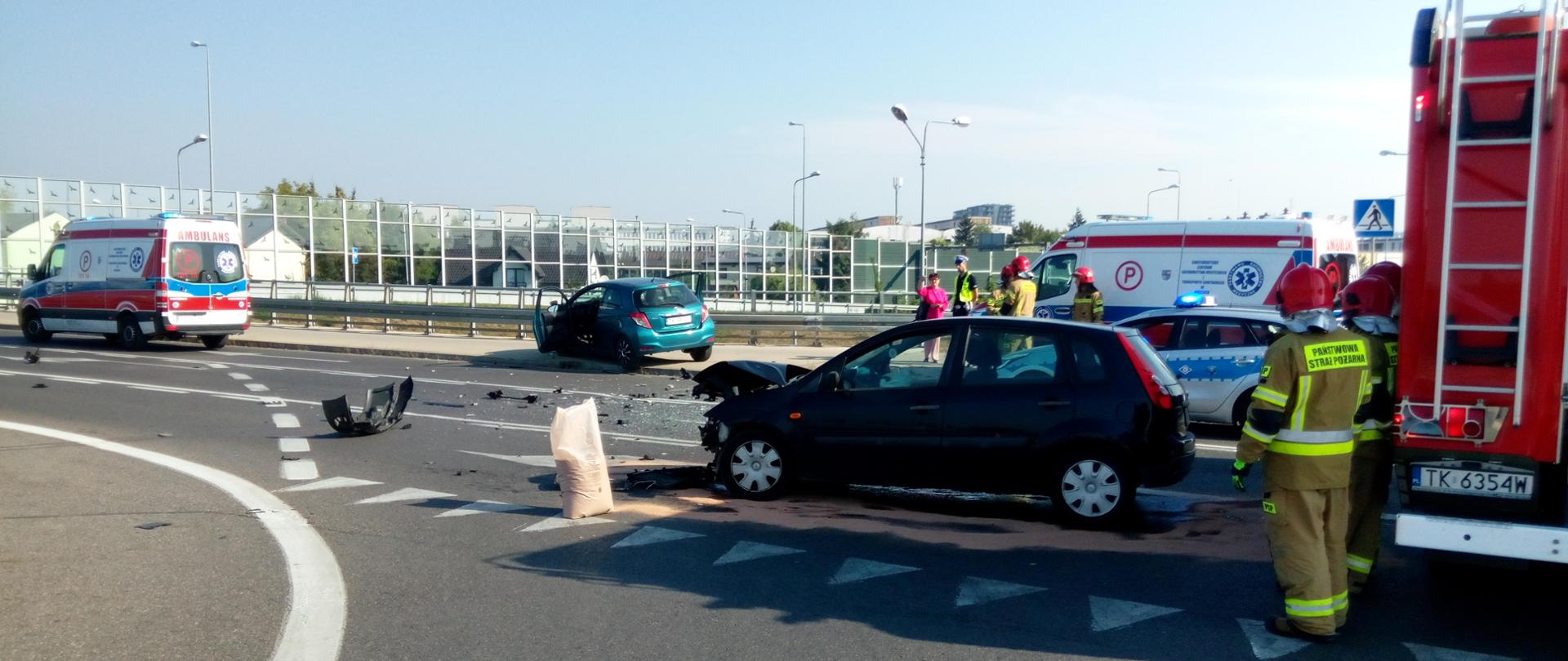 Zdjęcie przedstawia ulicę 1 Maja biegnącą górą wiaduktu. Od lewej widać stojący ambulans, dwa auta biorące udział w zderzeniu, w głębi drugi ambulans i radiowóz. Najbliżej widać ratownika stojącego przy wozie strażackim. Na jezdni znajduje się worek z sorbentem, którego znaczna ilość rozsypana jest na jezdni, leżą także rozrzucone fragmenty pojazdów.