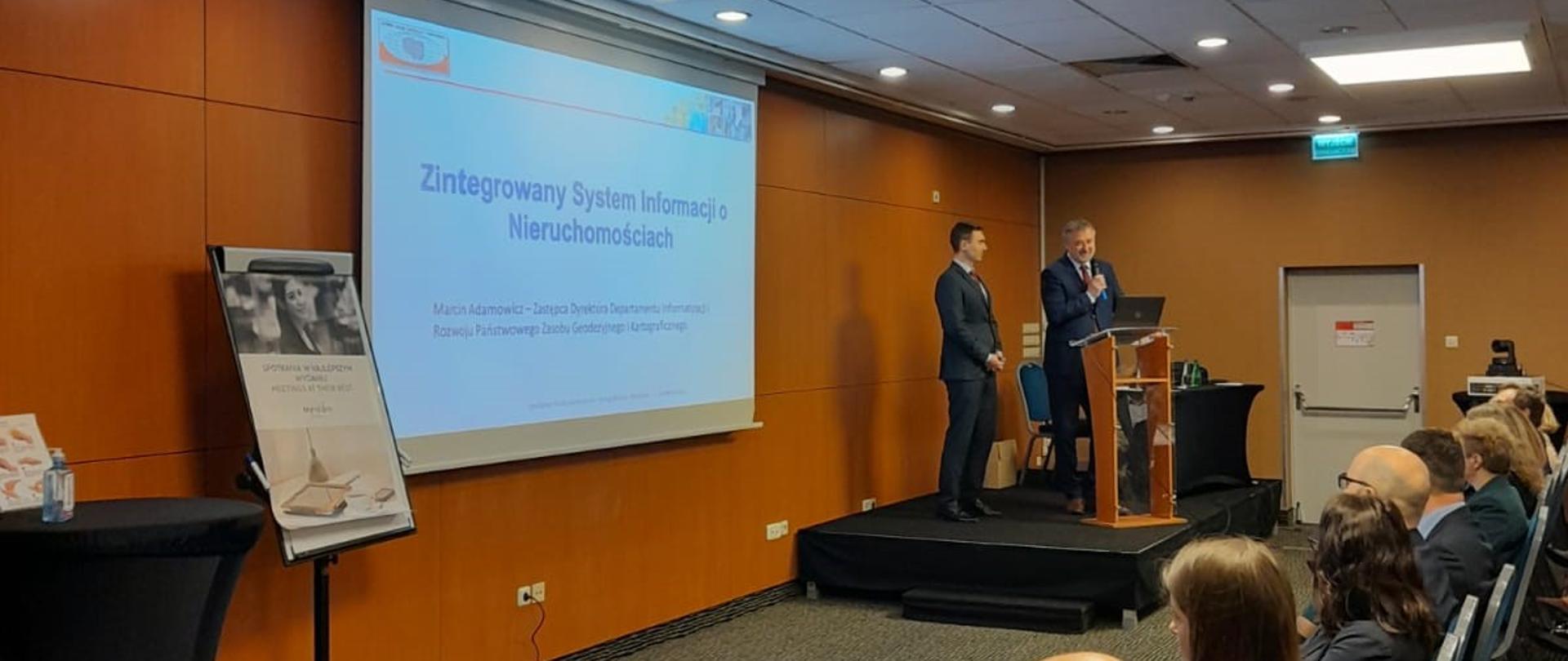 Zdjęcie przedstawia Głównego Geodetę Kraju dr. hab. inż. Waldemara Izdebski oraz Zastępcę Dyrektora Departamentu Departamentu Informatyzacji i Rozwoju Państwowego Zasobu Geodezyjnego i Kartograficznego - Marcina Adamowicza prezentującego informacje na temat ZSIN.