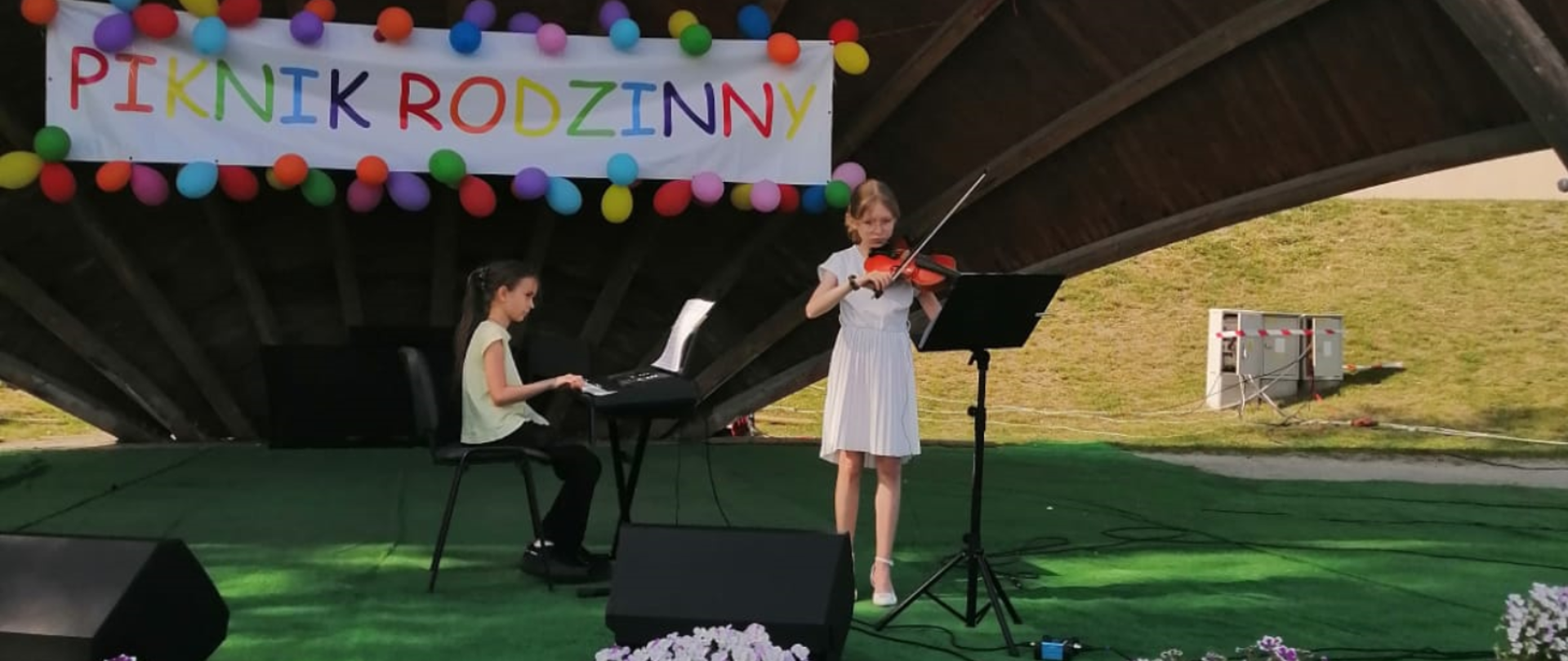 Na zdjęciu dwie dziewczyny. Jedna w ciemnych włosach, białej bluzce i czarnych spodniach siedzi na krześle i gra na keybordzie. Druga dziewczyna to blondynka w okularach i białej sukience, gra na skrzypcach. Przed nią stoi czarny pulpit . W tle na białym tle kolorowy napis: "piknik rodzinny". Dokoła napisu kolorowe balony. podłoże sceny w kolorze zielonym. 