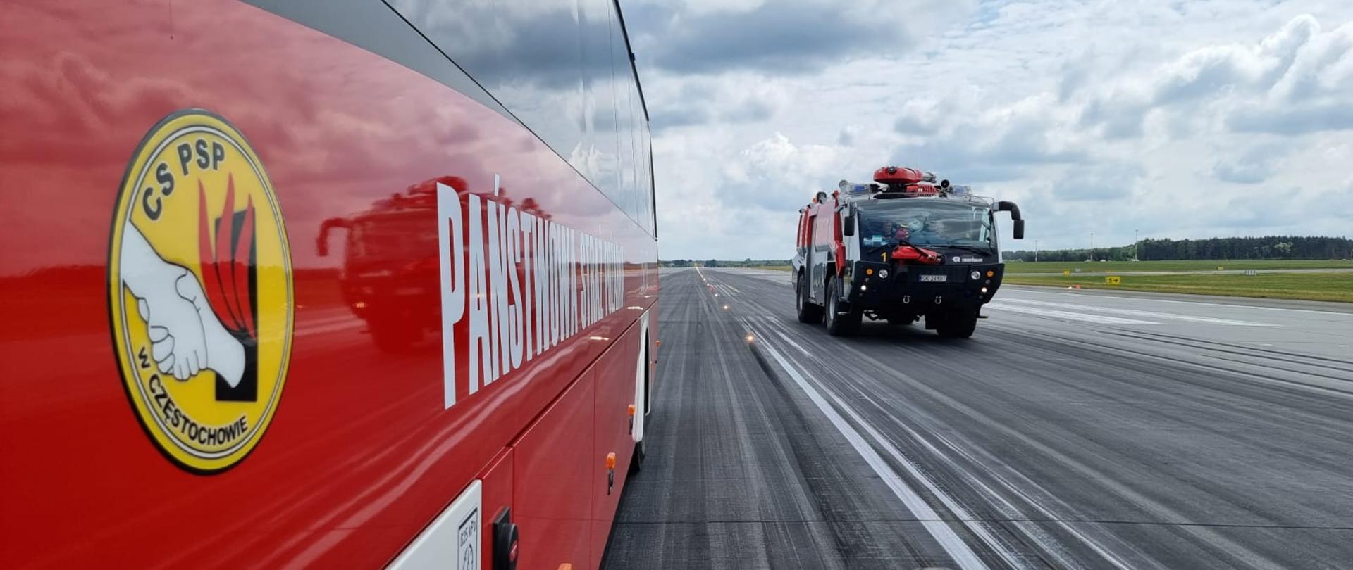 W głębi po prawej lotniskowy pojazd pożarniczy jadący o płycie lotniska po lewej widoczny bok autobusu z logo CS PSP