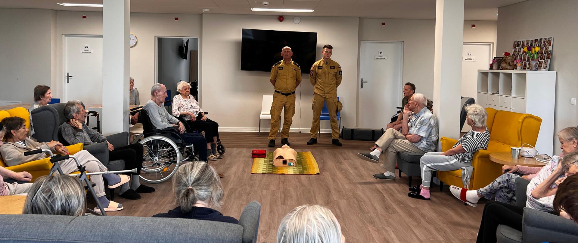 Na zdjęciu widać dwóch funkcjonariuszy, ubranych w piaskowe ubrania, którzy stoją na środku pomieszczenia. Przed nim leży fantom osoby dorosłej do resuscytacji, za nimi wisi telewizor. Wokół funkcjonariuszy siedzą osoby starsze na fotelach oraz wózkach inwalidzkich.
