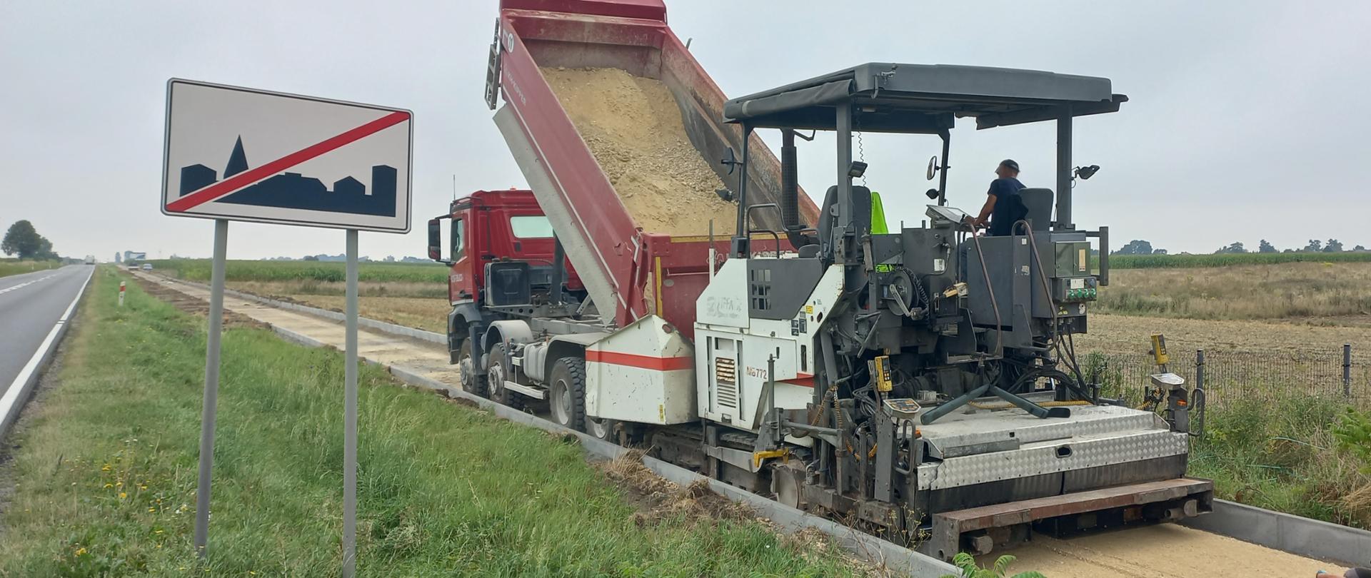 DK78 budowa ścieżki pieszo-rowerowej - jednojezdniowa droga asfaltowa, wzdłuż drogi budowany ciąg pieszo-rowerowy, na ścieżce pracuje układarka kruszywa do wykonywania podbudowy bitumicznej, przed nią czerwona ciężarówka-wywrotka z kruszywem, przy drodze tablica oznaczająca koniec terenu zabudowanego. Drogę od ścieżki oddziała pas zieleni 