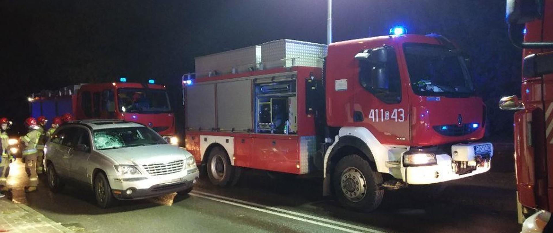 Na ulicy stoi srebrny samochód z widocznymi uszkodzeniami szyby po potrącaniu pieszego. Na drugim planie samochody strażackie w włączonymi sygnałami świetlnymi oraz najaśnicami oświetlającymi miejsce zdarzenia. W tle strażacy biorący udział w działaniach.