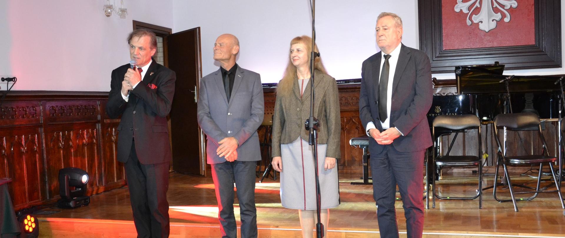 Cztery osoby stoją na scenie podczas Koncertu Patriotycznego w Sali Królewskiej, jedna z nich przemawia do mikrofonu podczas uroczystego wręczenia medali i nagród.