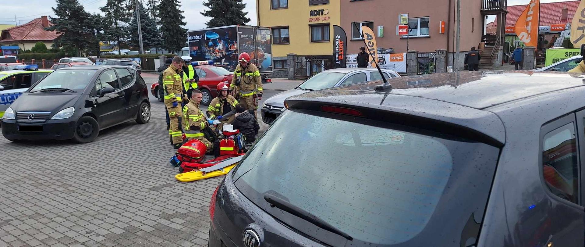 Parking przed sklepem. Na placu przed sklepem. Pomiędzy zaparkowanymi autami osobowymi strażacy udzielaja kwalifikowanej pierwszej pomocy potrąconej kobiecie. Poszkodowana znajduje się w pozycji siedzącej. Obok leżą nosze typu deska oraz torby ze sprzętem do ratownictwa medycznego. W tle droga, po której poruszaja sie pojazdy oraz budynki. 