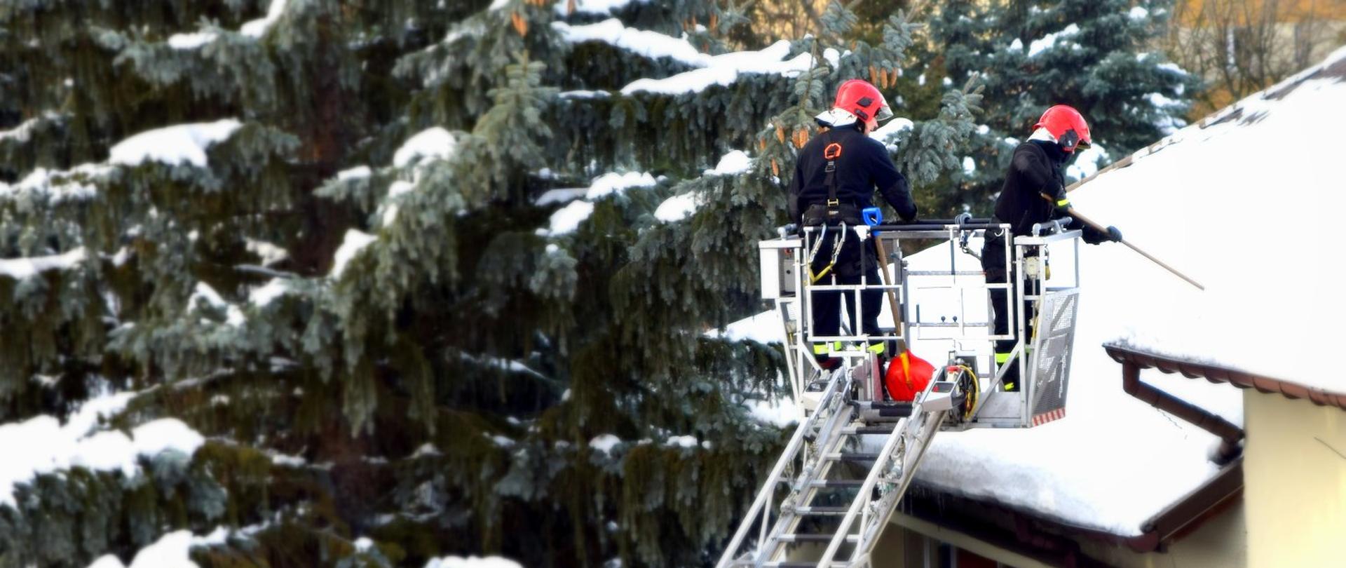 Zdjęcie zrobione na zewnątrz. Dwóch strażaków, będących w koszu drabiny mechanicznej, usuwa śnieg z dachu.