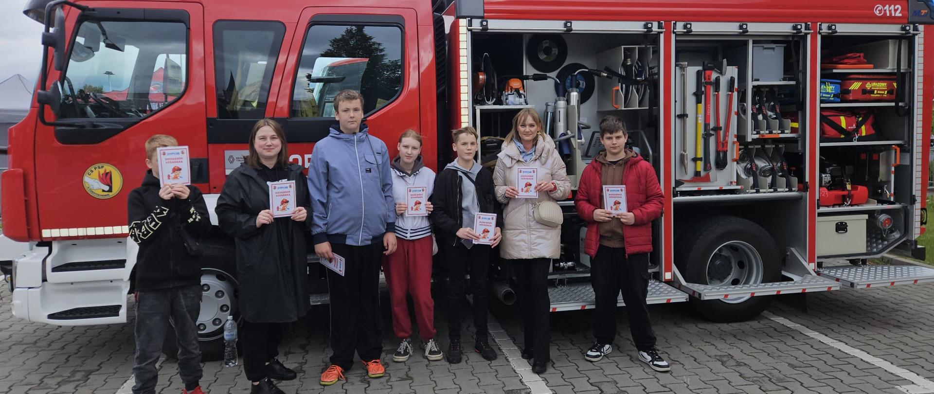 Grupa dzieci trzymająca w dłoniach pamiątkowe dyplomy stojąca na tle samochodu bojowego CS PSP, samochód ma otwarte schowki narzędziowe