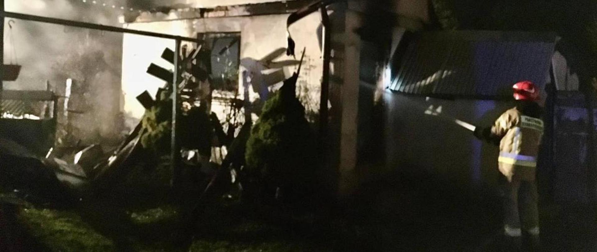 Zdjęcie przedstawia strażaka w ubraniu bojowym podczas gaszenia wodą objętego pożarem budynku gospodarczego. Zdjęcie wykonano w nocy.