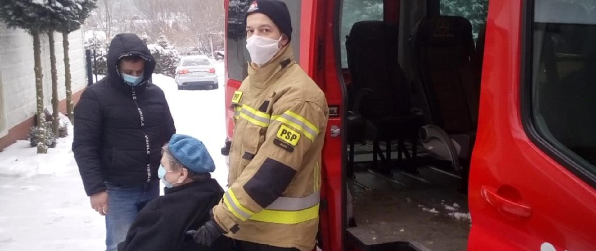 Na zdjęciu strażak z Jednostki Ratowniczo-gaśniczej w Żarach pomaga osobie na wózku inwalidzkim dotrzeć do punktu szczepień przeciw Covid-19. Z lewej strony zdjęcia znajduje się czerwony samochód strażacki typu bus. 